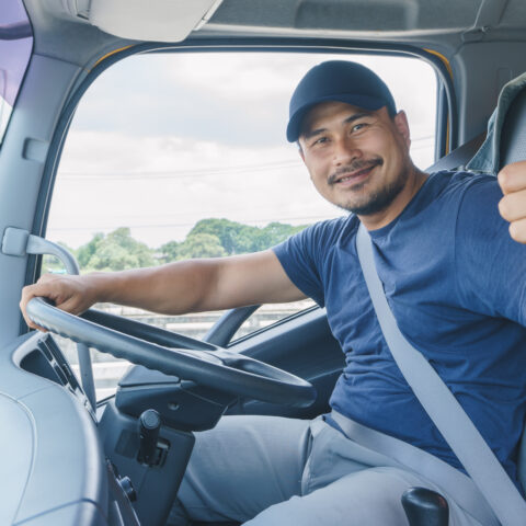 Young professional truck driver sat in the driver's seat wearing a navy t-shirt and cap, looking at the camera holding his thumb up