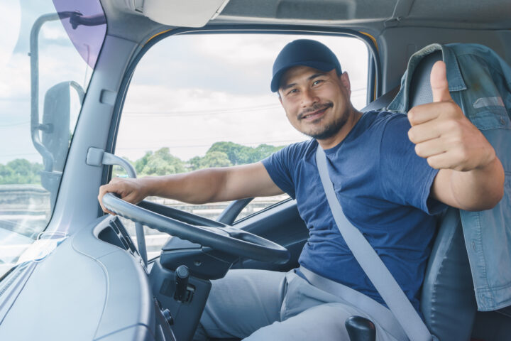 Young professional truck driver sat in the driver's seat wearing a navy t-shirt and cap, looking at the camera holding his thumb up